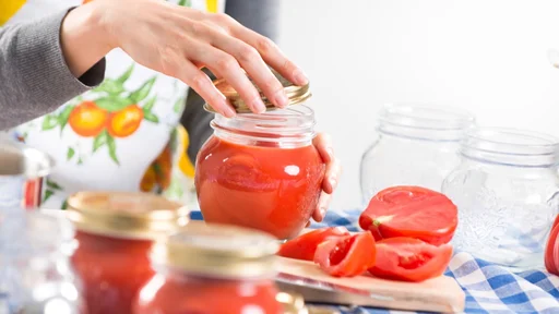 Woman filling glass jars with tomato sauce on checked tablecloth, hands close-up.