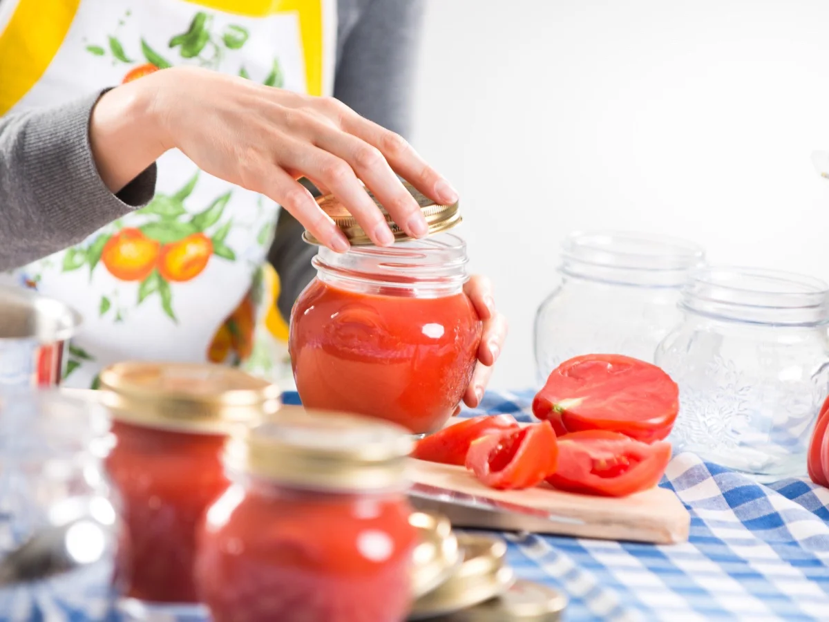Woman filling glass jars with tomato sauce on checked tablecloth, hands close-up.