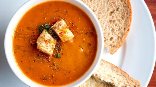 A bowl of fresh tomato soup in white ceramic bowl, garnished with herbs, croutons, seasoning and a drizzle of olive oil, and served with crusty wholemeal bread.