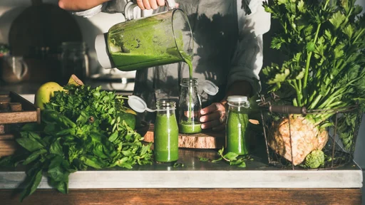 Making green detox take-away smoothie. Woman in linen apron pouring green smoothie drink from blender to bottle surrounded with vegetables and greens. Healthy, weight loss food concept (Making green detox take-away smoothie. Woman in linen apron pouri