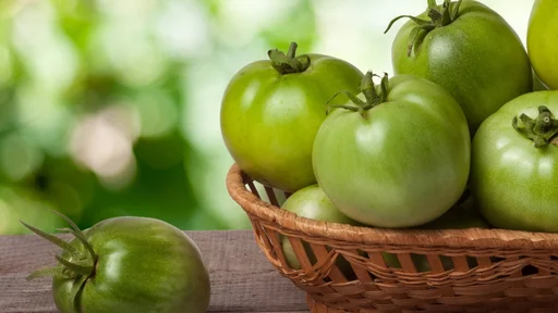unripe green tomatoes in a wicker basket on a wooden table with a blurred background.