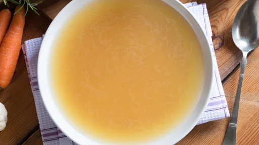 Bone broth made from chicken on a wooden table, with vegetables in the background, top view