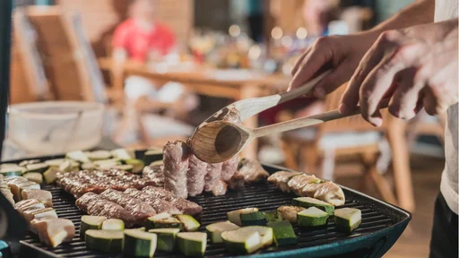Hands of a man seen flipping cevapcici meal on a grill at an evening party setting. Other guests waiting for their meal in the background.