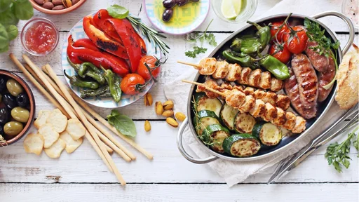 Grilled meat, chicken skewers and sausage with roasted vegetables and appetizers variety serving on party outdoor table. Mediterranean dinner table concept. Overhead view.