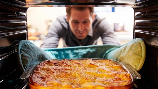 Man Taking Cooked Dish Of Lasagne Out Of The Oven. Smiling