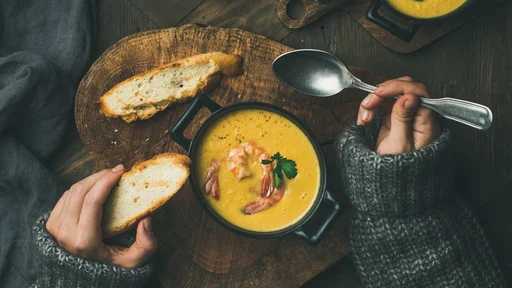 Woman in warm grey sweater eating corn creamy chowder soup with prawns served in individual pots, top view. Woman' s hand keeping spoon and bread slice. Flat-lay of rustic dinner table. Slow food, winter warming food concept