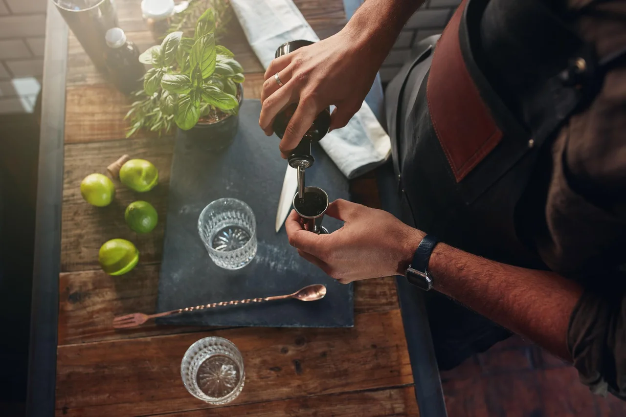Close up of barman hands pouring alcoholic drink in to a jigger to prepare a cocktail, with basil leaves and lemons on the bar counter.