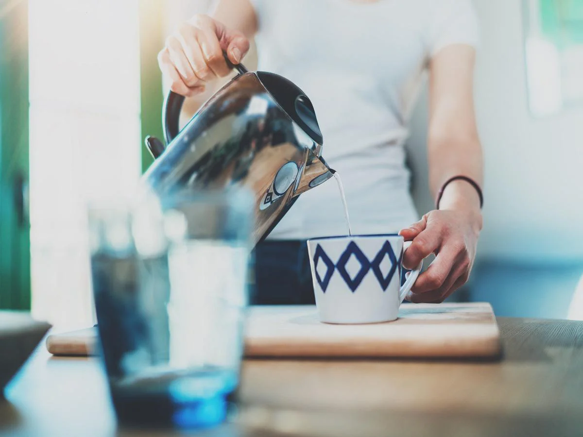 Young woman using kettle for make tea or black coffee on kitchen at room interior background.Women's hands pour water from a teapot into a cup. Blurred, flares effect