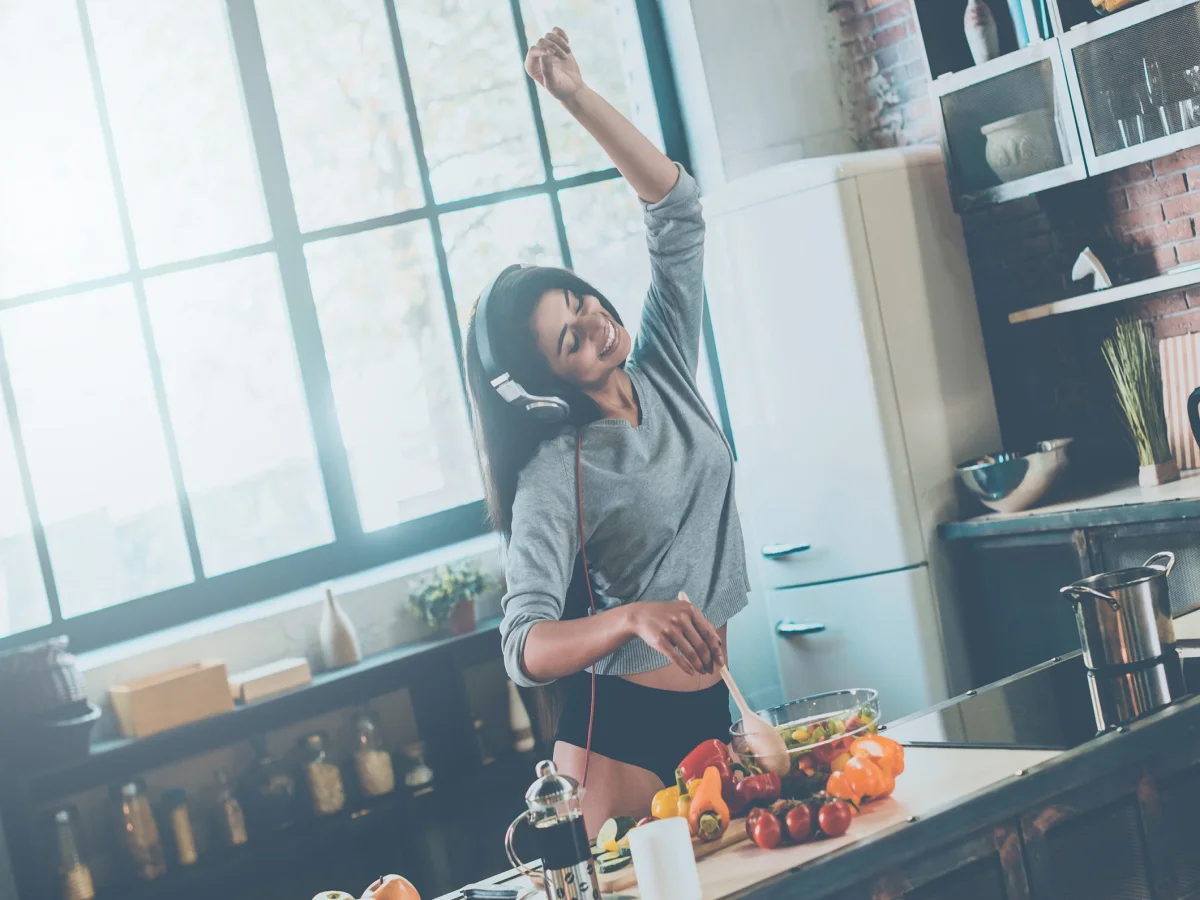 Beautiful young mixed race woman in headphones cooking salad and dancing while standing in kitchen at home