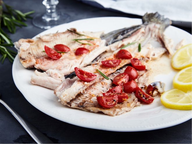 Delicious baked trout with cherry tomatoes, olive oil, lemon juice and fresh rosemary on white plate with lemon slices and fork. Behind blurred glass of red wine, rosemary and towel. Black background (Delicious baked trout with cherry tomatoes, olive